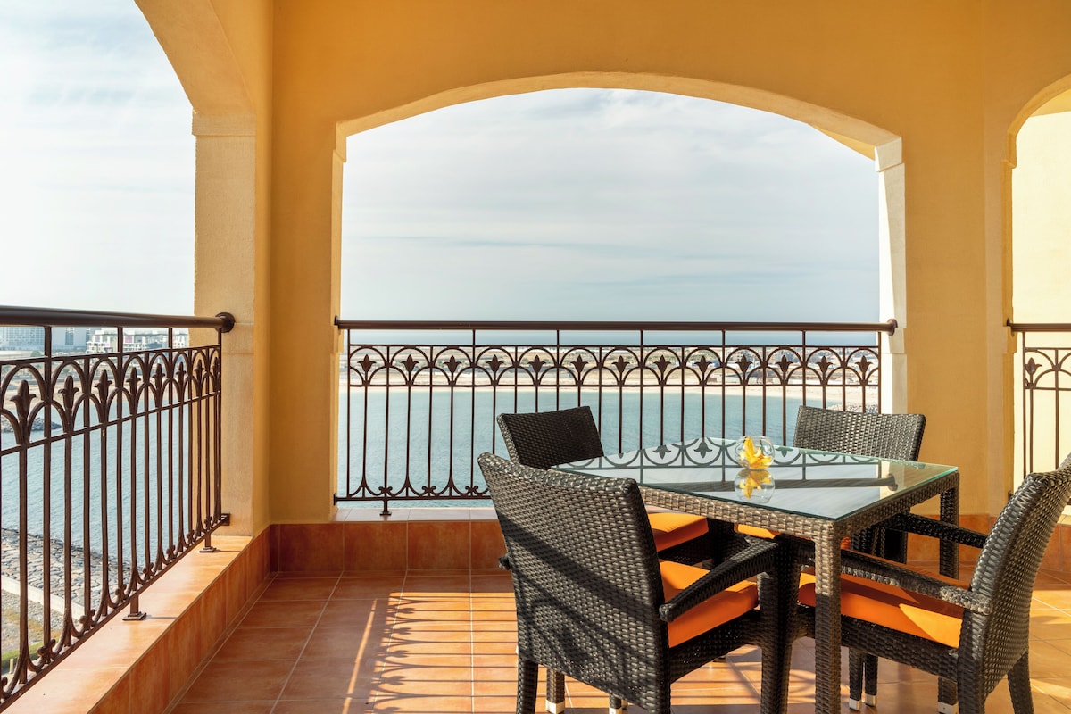 A balcony with a glass table and wicker chairs, overlooking a body of water.
