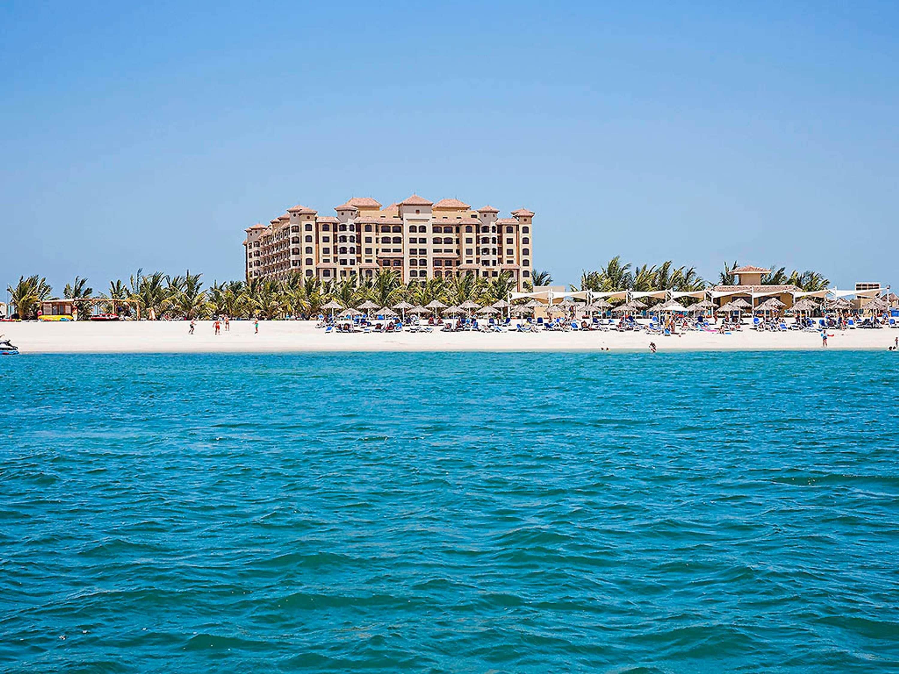 Ein Strandhotel mit mehreren Balkonen, einem Sandstrand und einem klaren, blauen Meer.