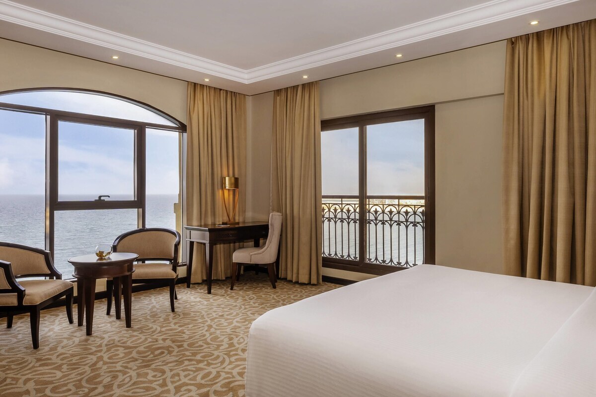 Habitación de hotel con una cama grande, un escritorio con silla, una mesita y vistas al mar a través de amplios ventanales.
