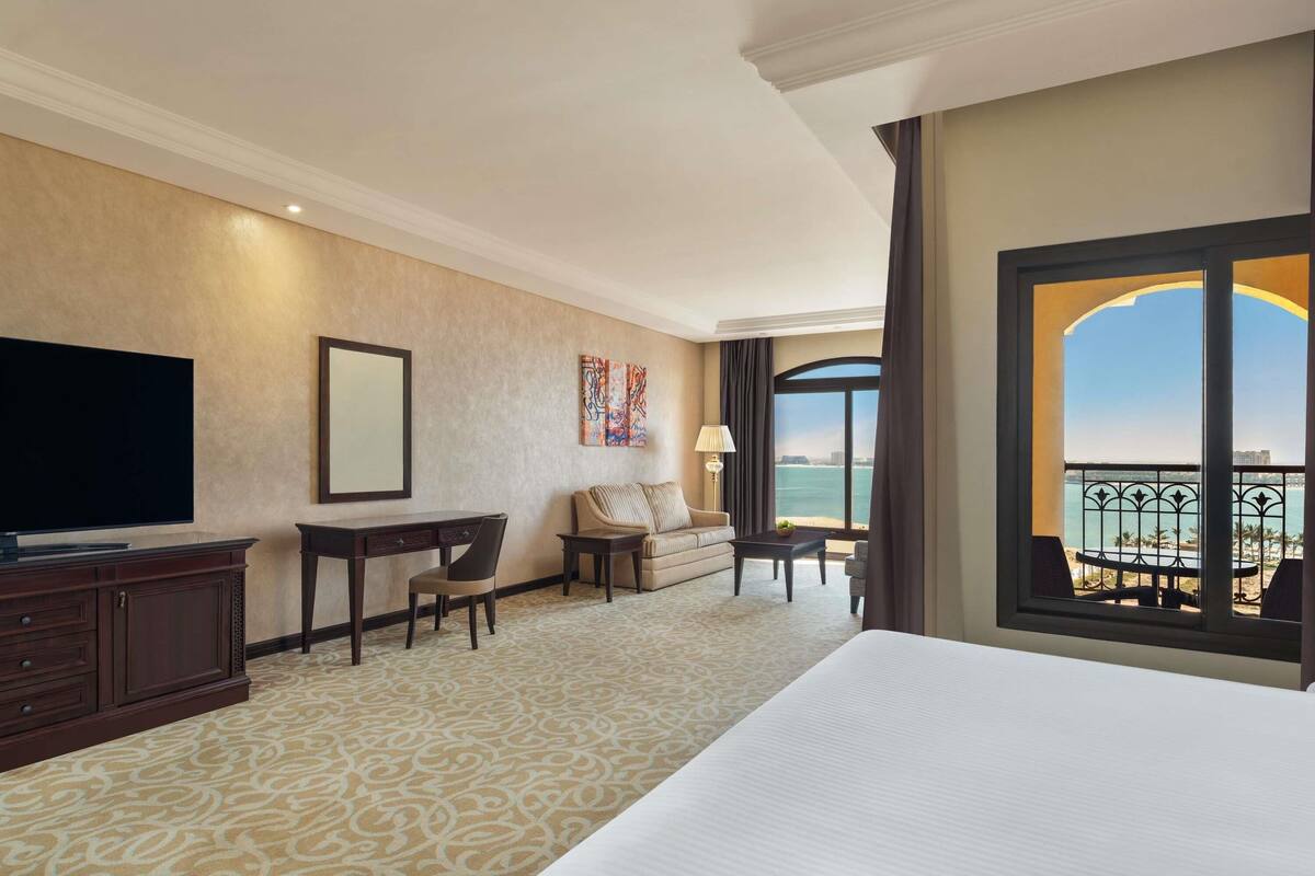 Une chambre d’hôtel avec un grand lit, un bureau, une chaise, une télévision et une vue sur la mer depuis un balcon.