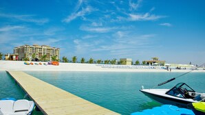 A marina with a wooden dock, a boat, and a beach with sun loungers and a hotel in the background.