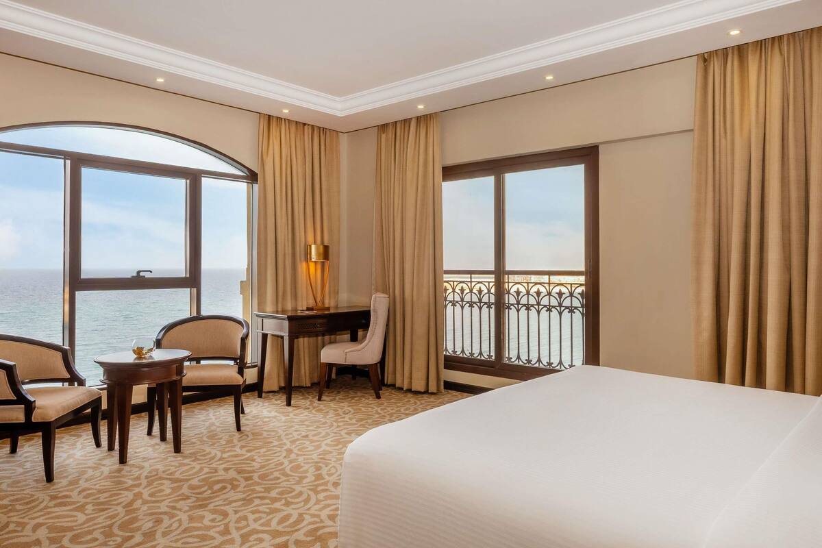 Une chambre d’hôtel dotée d’un grand lit, d’un bureau avec une chaise, d’une petite table avec une chaise, et offrant une vue sur la mer grâce à de grandes fenêtres.