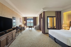 A hotel room with a large bed, a TV, a desk with a chair, a small table, and a view of the city through large windows. - Pullman Resort Al Marjan Island (Ras Al Khaimah)