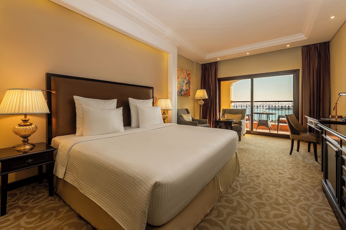 A hotel room with a large bed, a desk, a chair, and a balcony with a view.