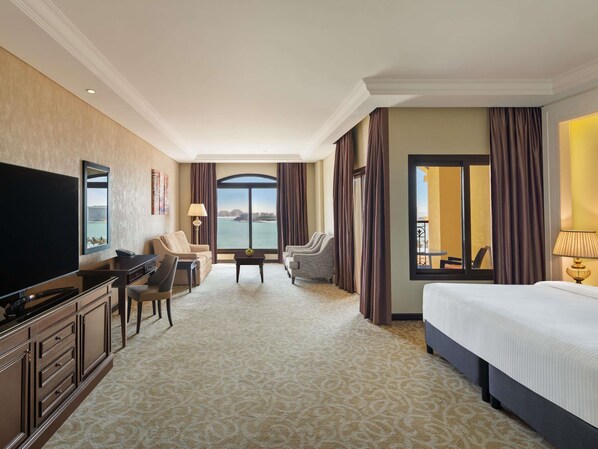 Habitación de hotel con una televisión grande, sofá, mesa de comedor, cama y vista al mar.
