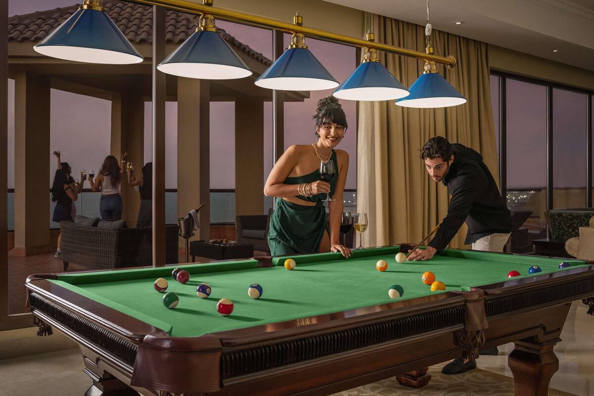 Une table de billard avec plusieurs billes, une personne qui joue et une autre qui se tient à côté. Il y a trois lampes bleues au-dessus de la table.