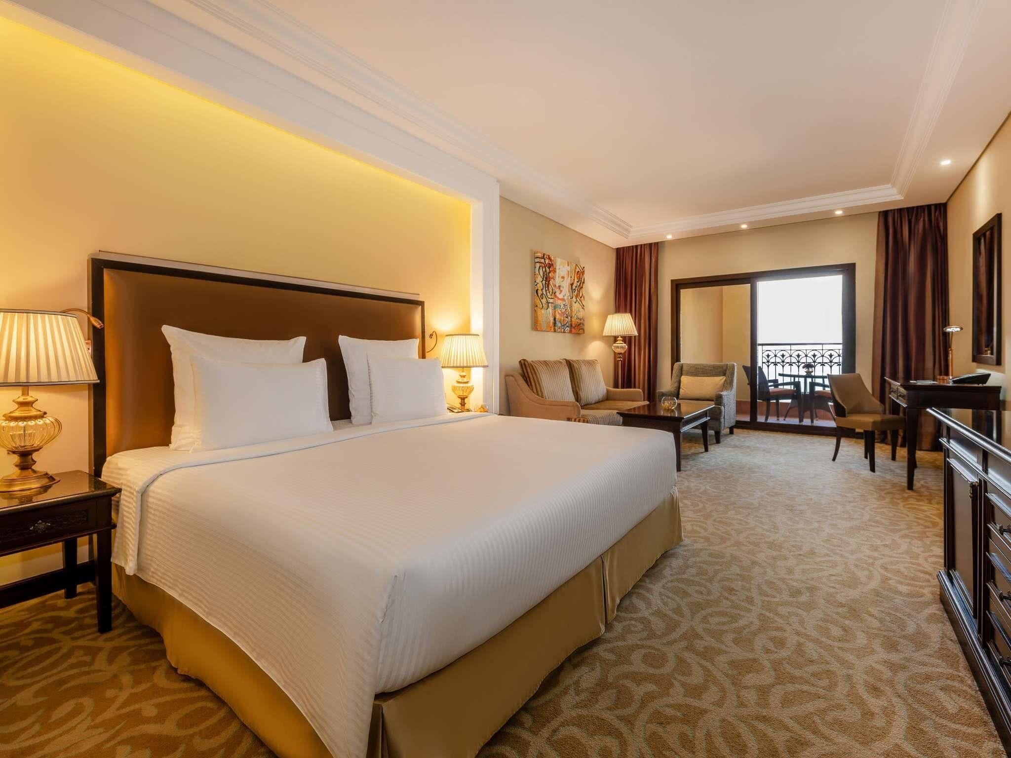 A hotel room with a large bed, a desk, a chair, and a balcony with a view.