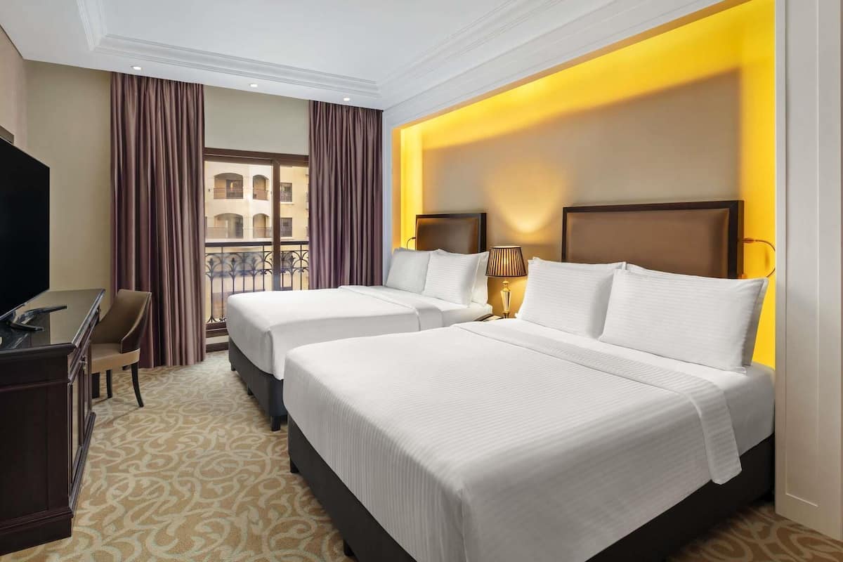 A hotel room with two beds, a desk, a TV, and a balcony with curtains.