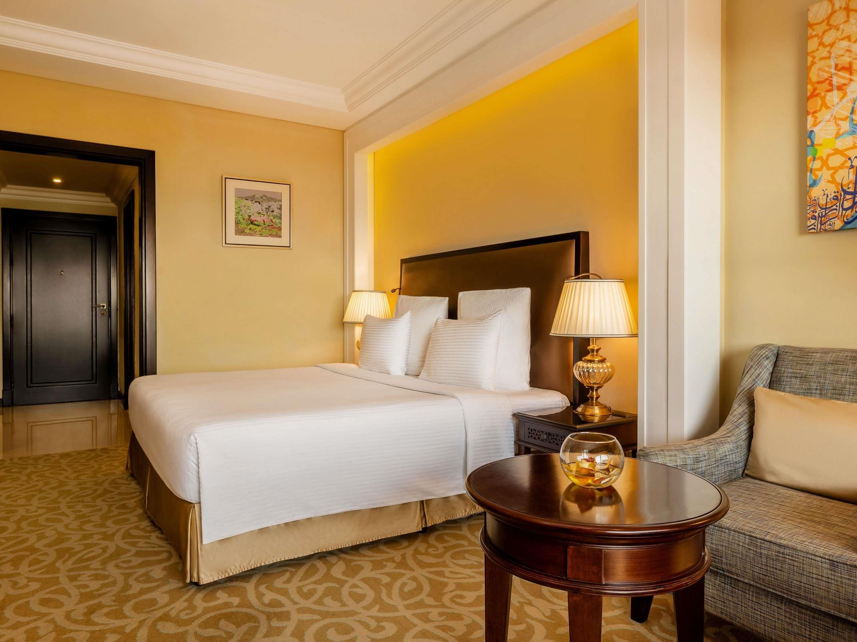 Quarto de hotel com uma cama grande, duas luminárias de cabeceira, uma mesinha com um copo e um sofá.