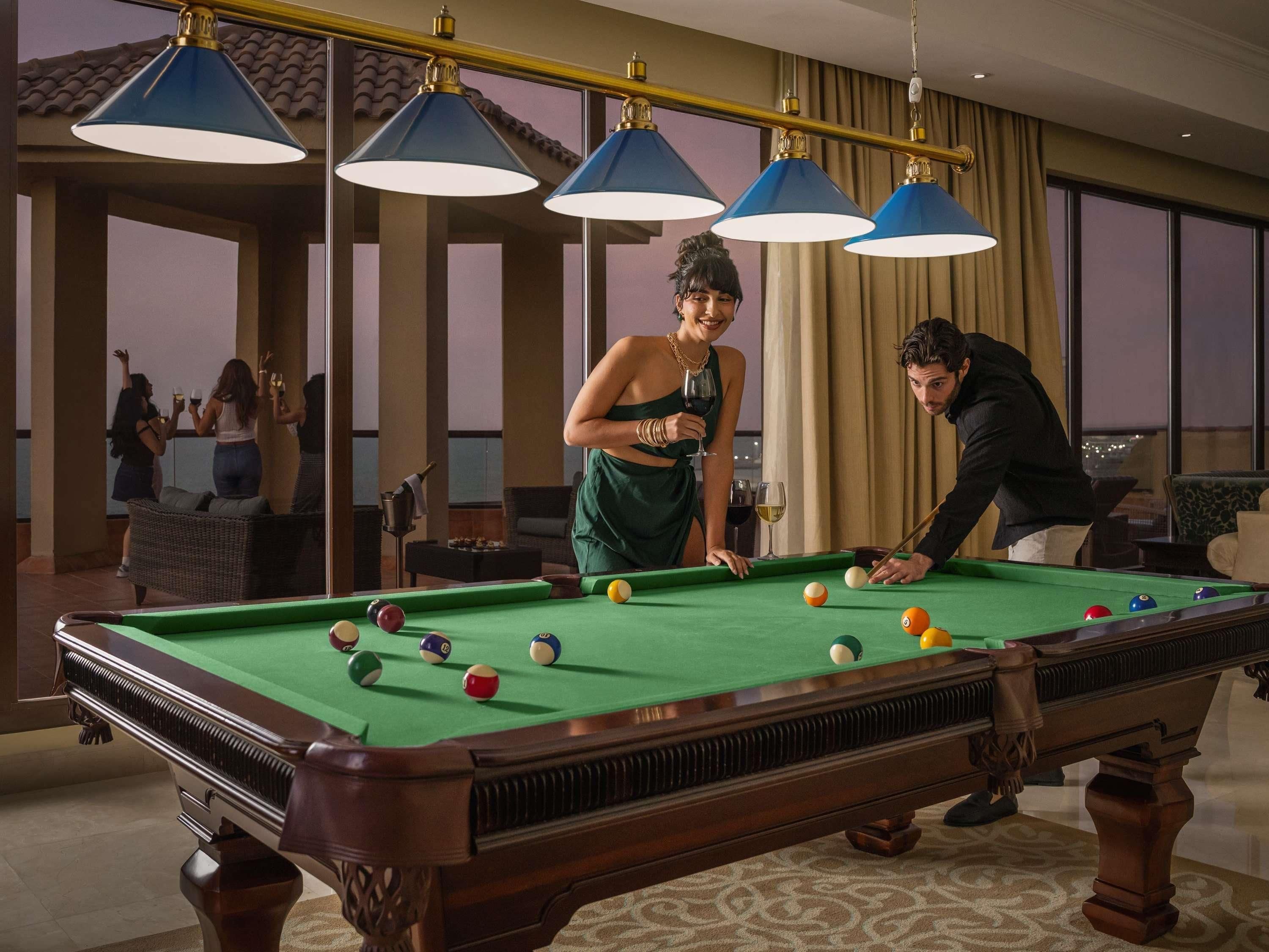 Une table de billard avec plusieurs billes, une personne qui joue et une autre qui se tient à côté. Il y a trois lampes bleues au-dessus de la table.