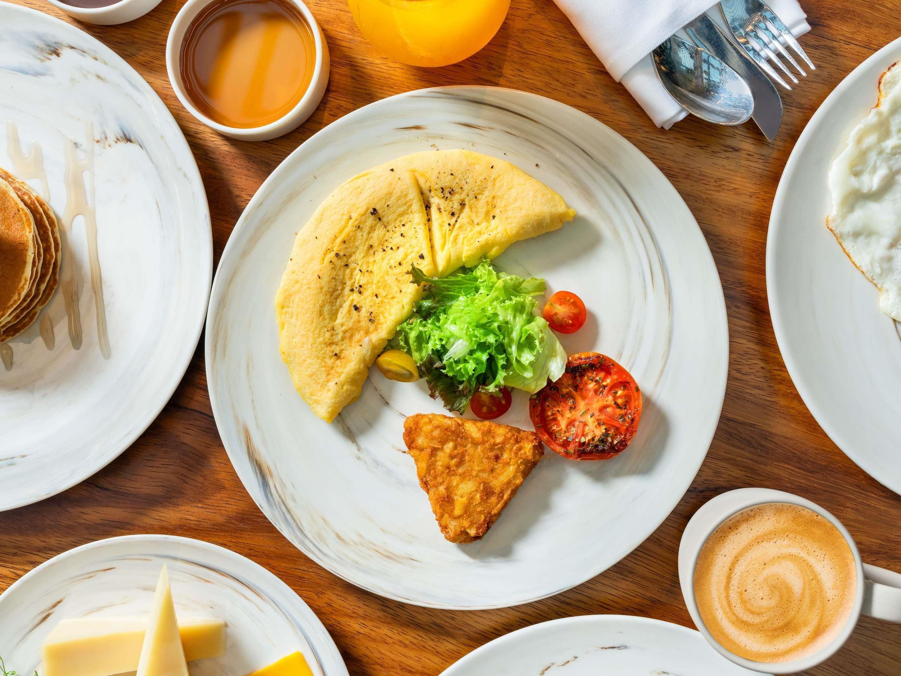 A breakfast setting with plates of food, including pancakes, an omelette, a fried item, and a cup of coffee.