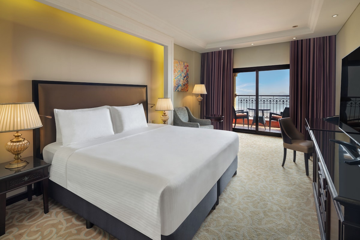 A hotel room with a large bed, a desk with a TV, a chair, and a balcony with outdoor seating.