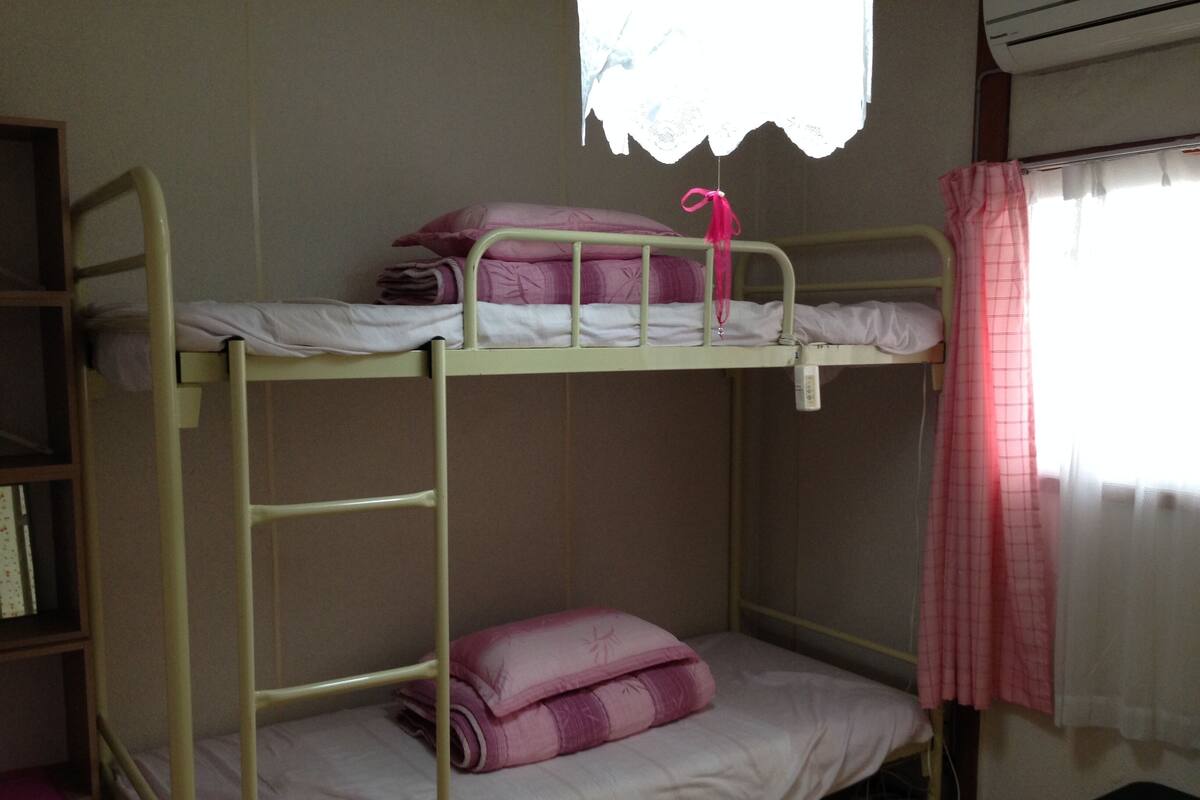 A bunk bed room with a ladder, a window with a lace curtain, and a bedside table with a pink item.