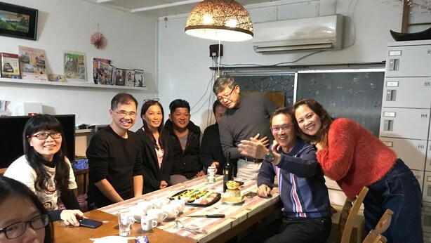 A group of people seated around a wooden table with food and drinks, in a room with white walls, a hanging lamp, and an air conditioning unit. - IM Guest House - Hostel (Osaka)