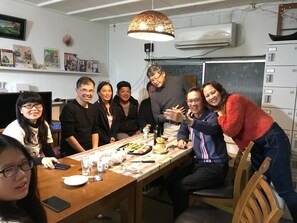 A group of people seated around a wooden table with food and drinks, in a room with white walls, a hanging lamp, and an air conditioning unit. - IM Guest House - Hostel (Osaka)