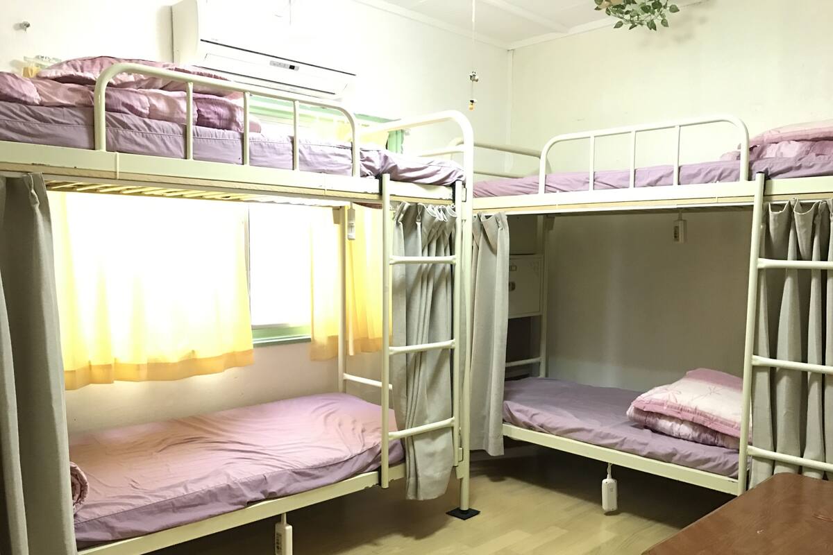 A dormitory room with bunk beds, a bedside table, and a window with curtains.