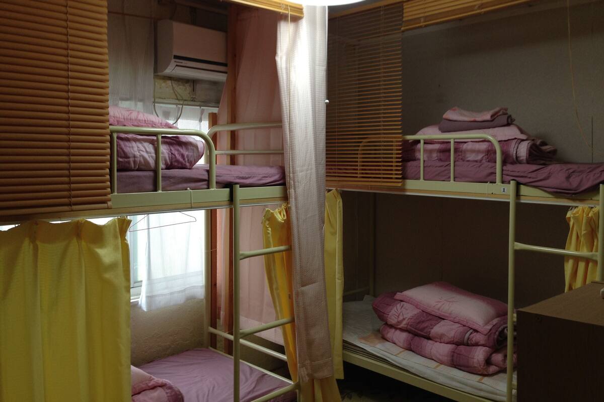 A dormitory room with bunk beds, a window with blinds, an air conditioning unit, and a hanging light fixture.