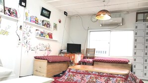 A compact room with a bed, a desk, a chair, and a TV. There are shelves with books and decorations, and an air conditioning unit on the wall. - IM Guest House - Hostel (Osaka)