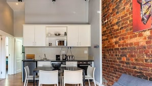 A modern kitchen with white cabinets, a dining area with a gray sofa, and a brick wall with a framed picture. - Long Street Boutique Hotel (Cape Town)
