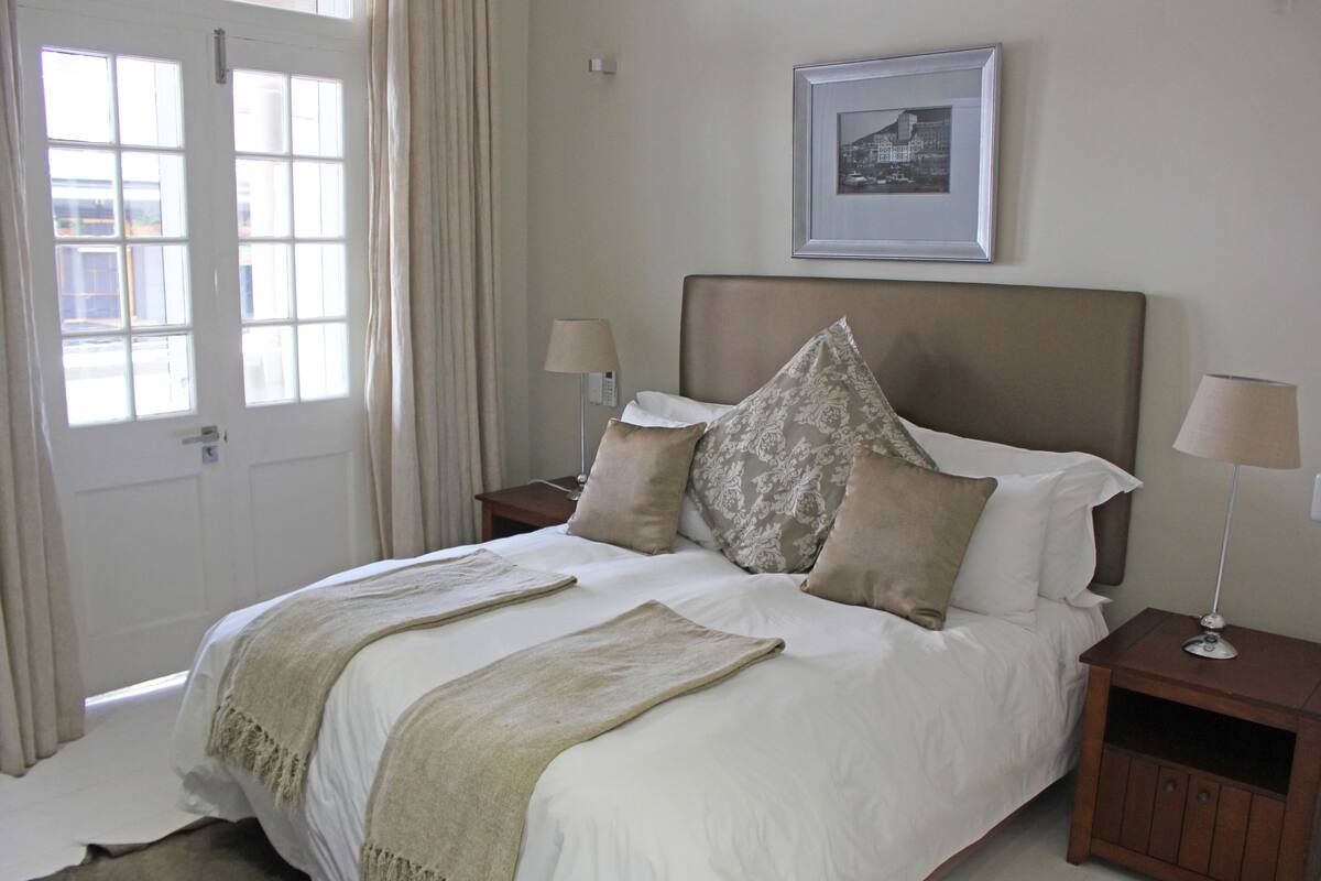 A neatly made bed with a headboard, two bedside tables with lamps, a framed picture on the wall, and a door with curtains.