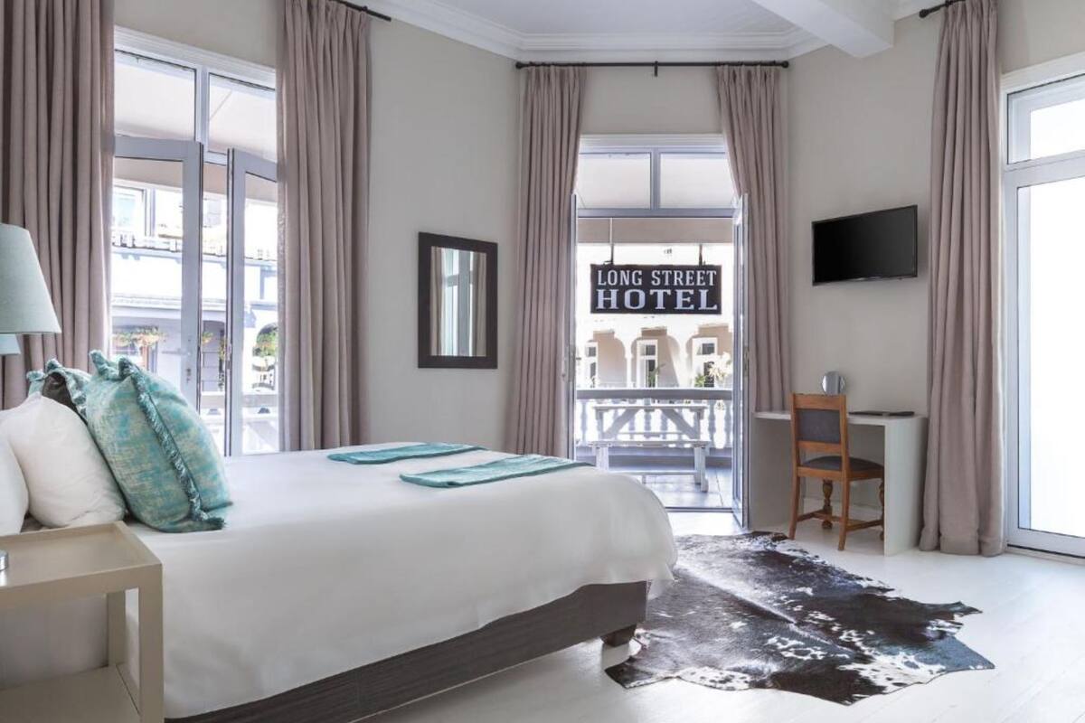 A hotel room with a large bed, a desk, a chair, a TV, and a view of the Long Street Hotel.