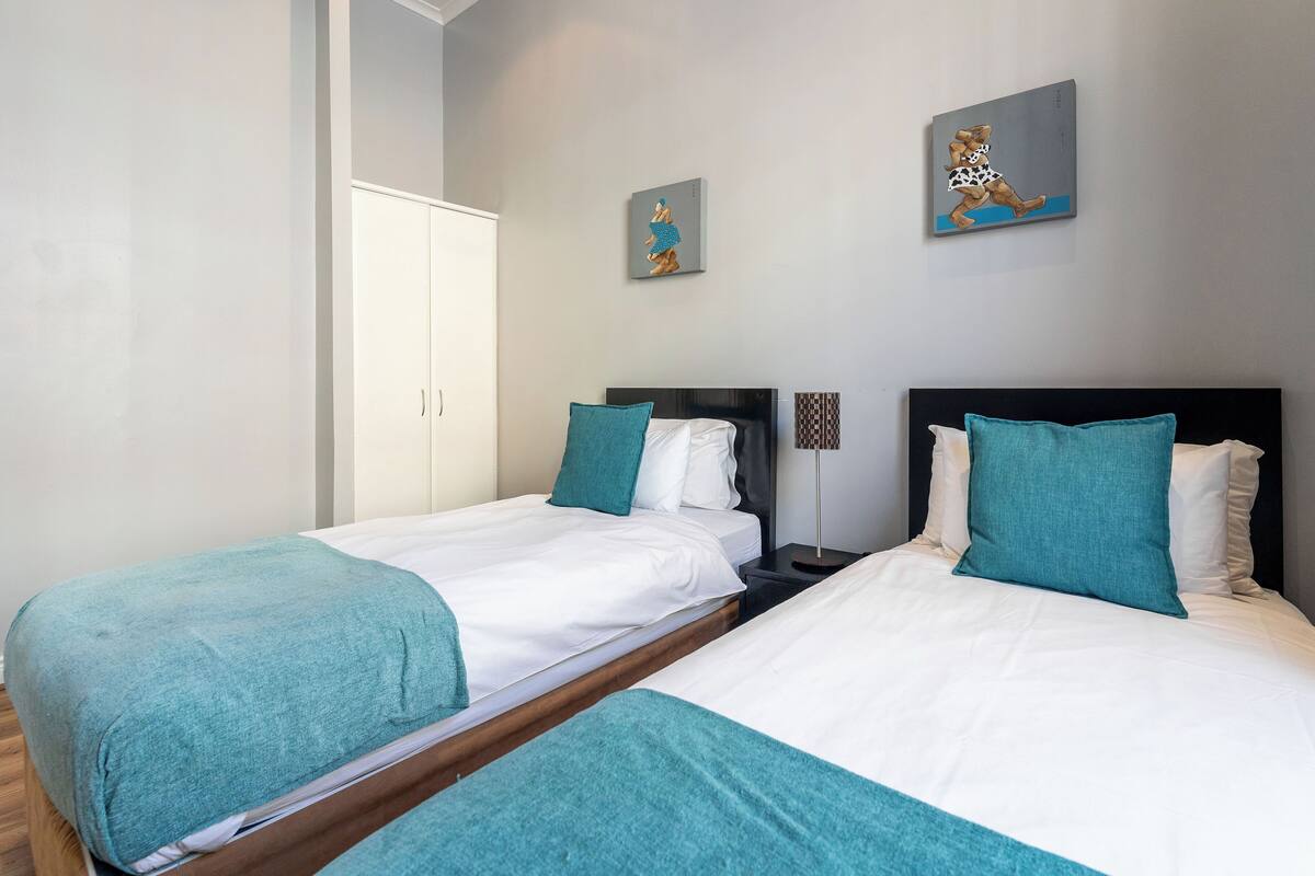 A hotel room with two beds, each with white linens and teal pillows. There are two paintings on the wall, a wardrobe, and a bedside table with a lamp.