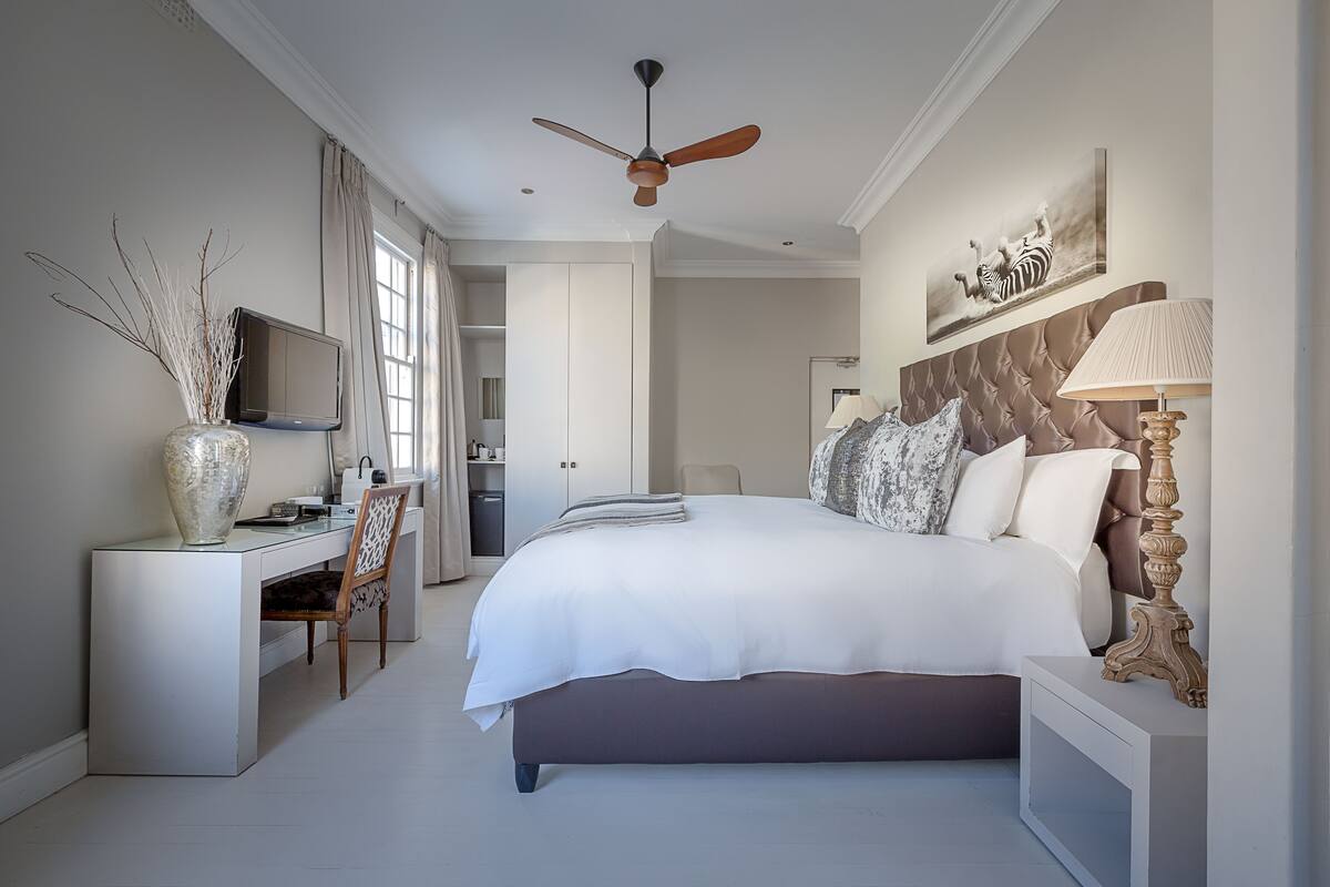 A bedroom with a large bed, a desk, a chair, a TV, and a ceiling fan.