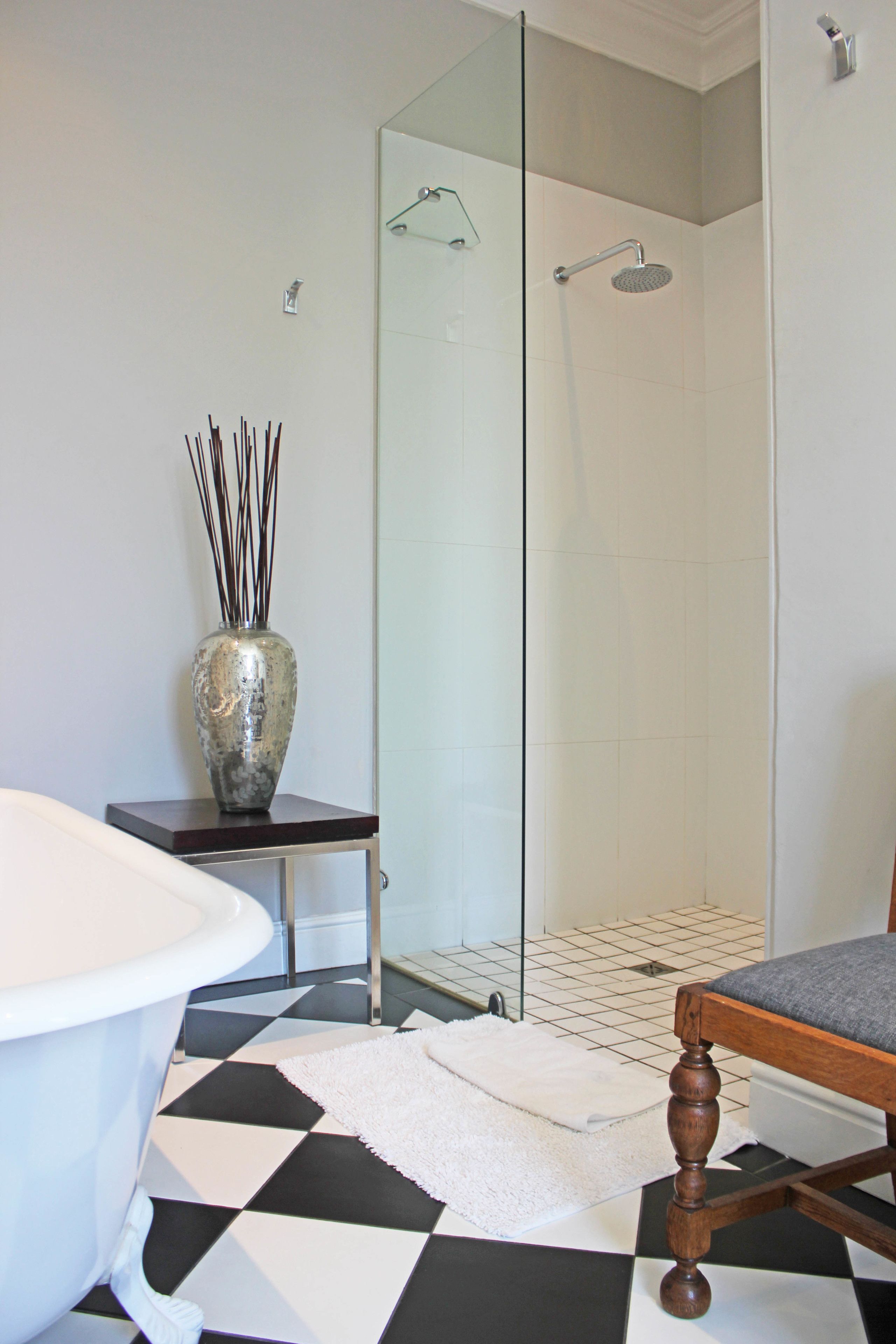 Une salle de bain avec une baignoire blanche, une douche à parois de verre, une chaise en bois et un vase contenant des roseaux, posé sur une petite table.