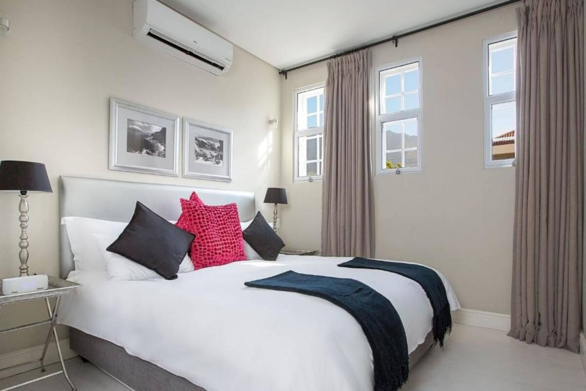 A hotel room with a large bed, two bedside tables, an air conditioning unit, and two windows with curtains.