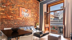 A modern living room with a brick wall, a dining area with a dark wooden table and white chairs, a grey sofa, and a large window with a view of buildings. - Long Street Boutique Hotel (Cape Town)