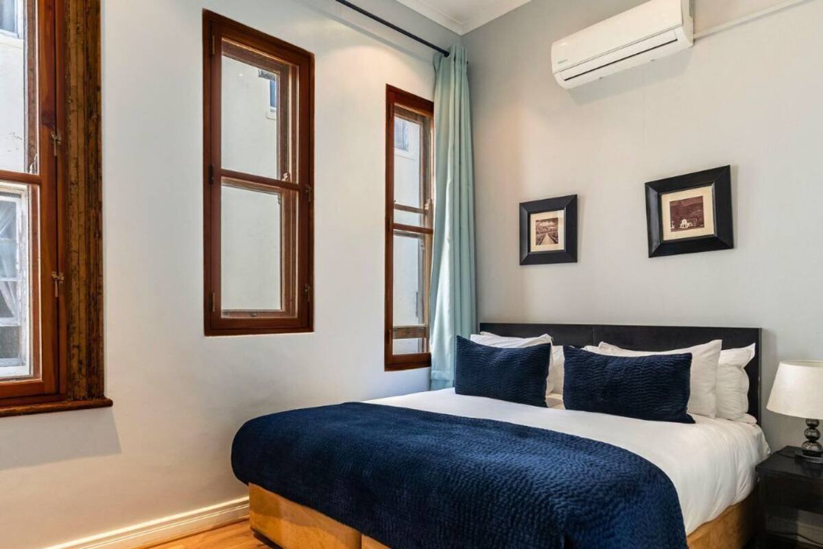A bedroom with a large bed, wooden floor, two windows, and a wall-mounted air conditioner.
