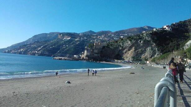 A coastal beach with a clear view of the sea, a sandy shore, and buildings on a hillside in the background. - B&B Vittoria (Maiori)