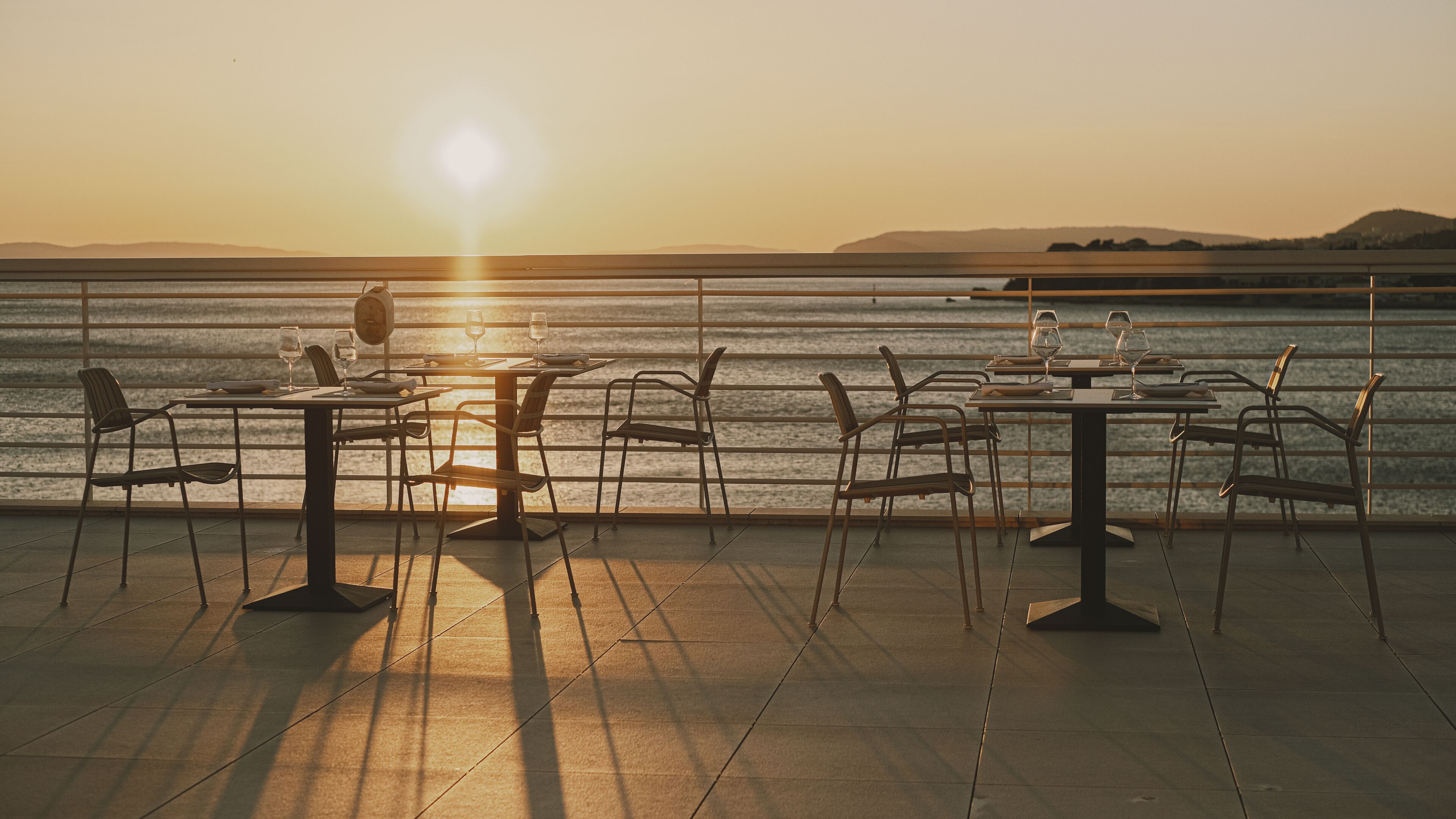 An outdoor dining area with tables and chairs facing a body of water during sunset.