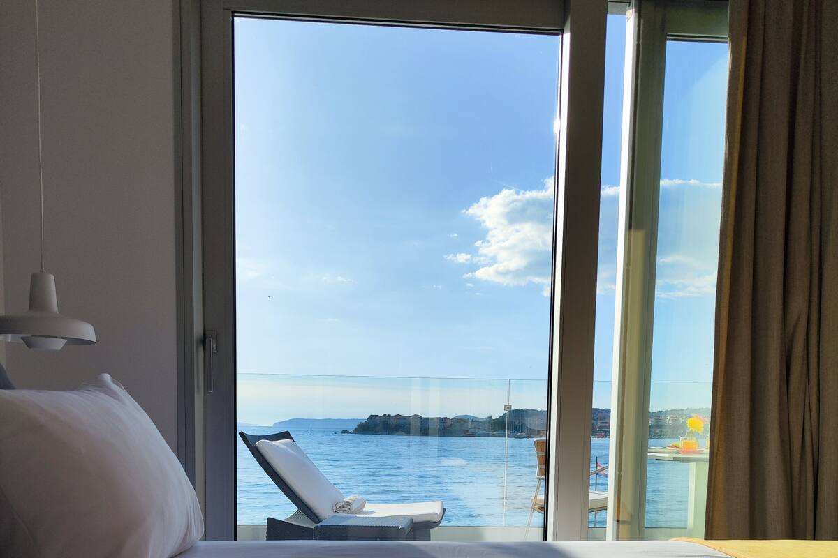 A hotel room with a bed covered in rose petals, a view of the sea, and a balcony with a chair.