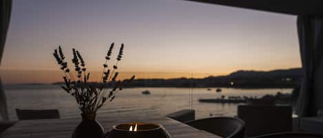 A table with a vase of flowers and a candle, overlooking a body of water with boats and a sunset view.