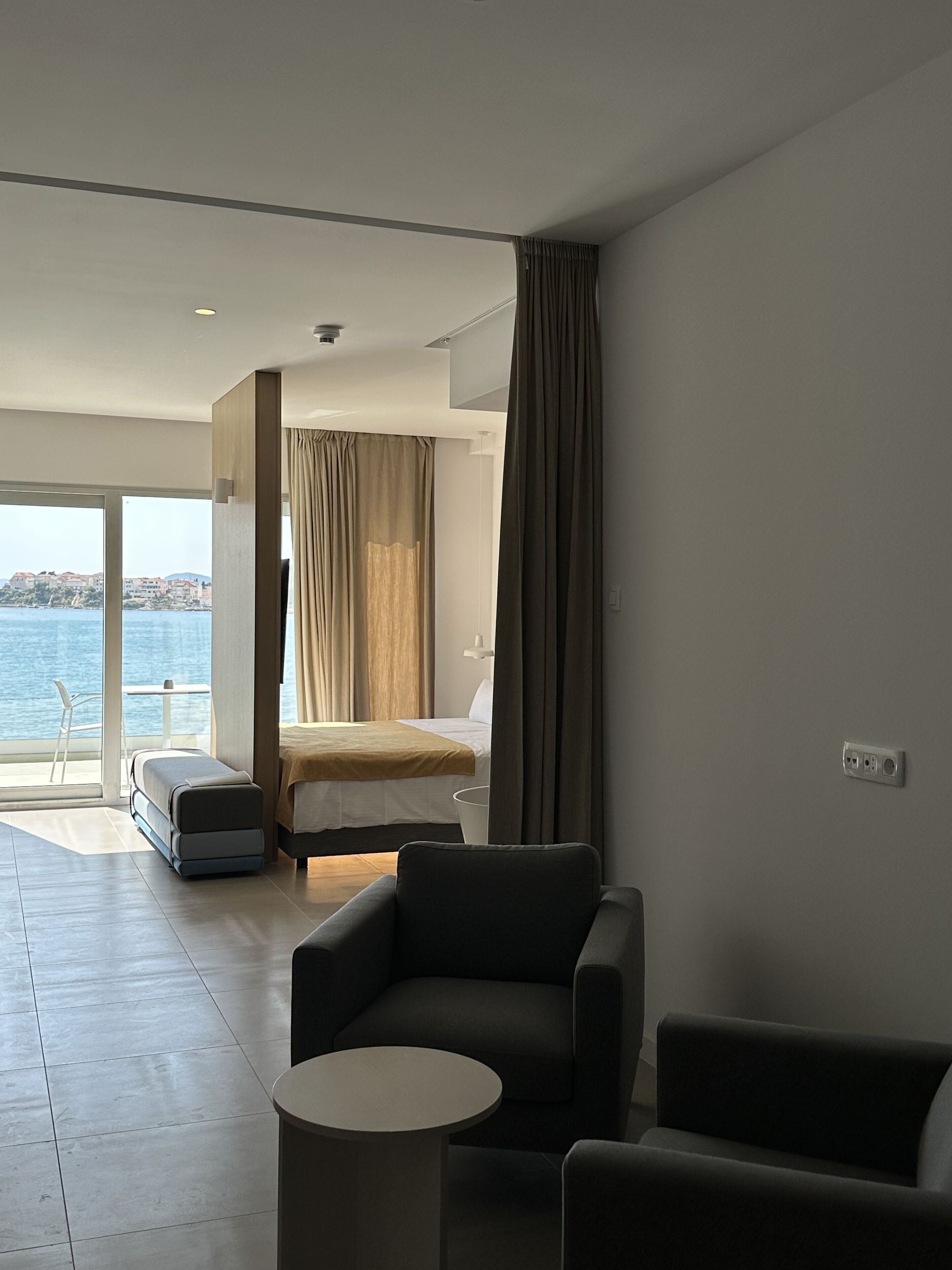 Ett modernt hotellrum med en säng, en sittgrupp med två fåtöljer och ett litet bord, en balkong med utsikt över havet och ett stort fönster.