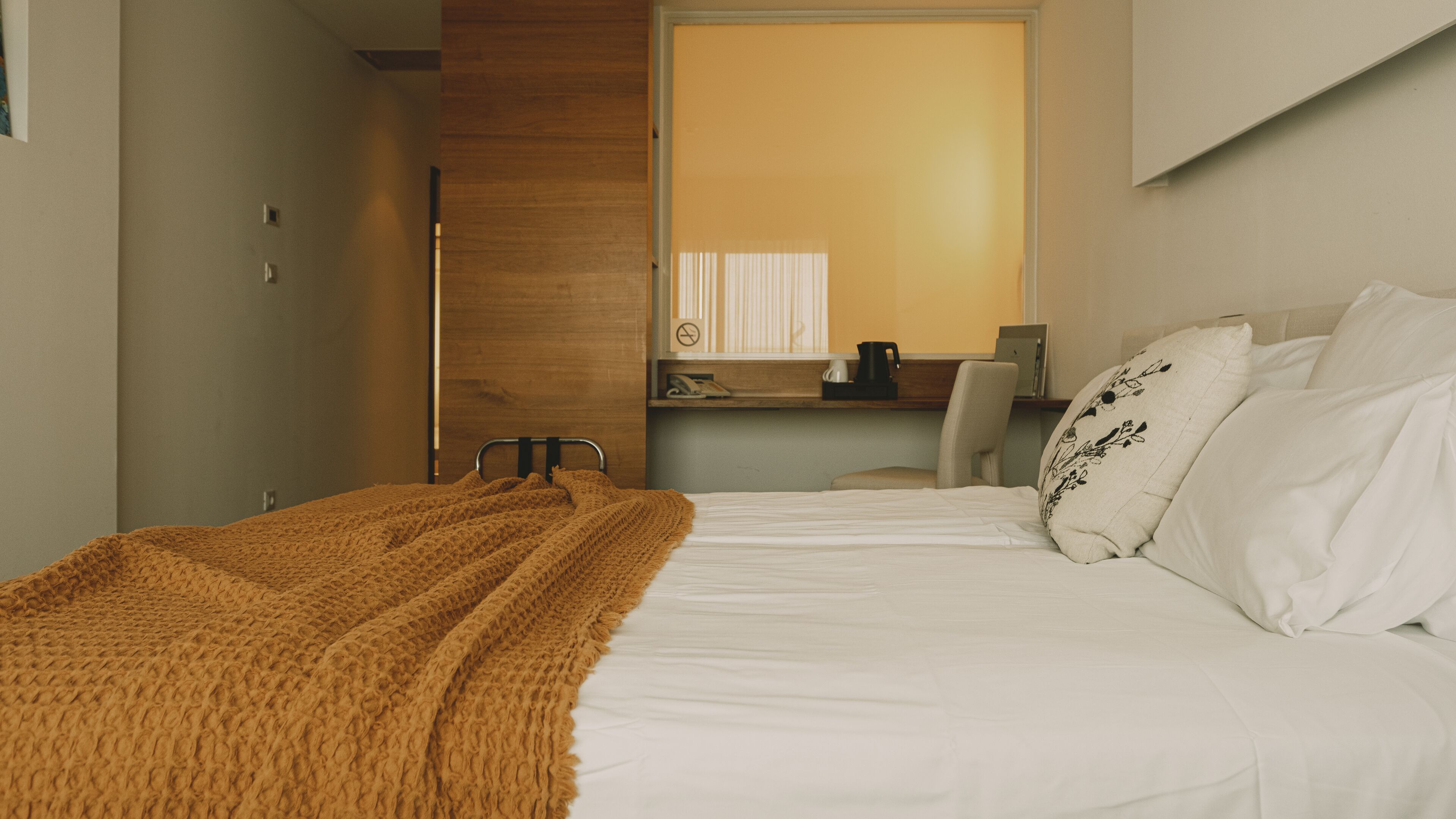 Una camera d'albergo con un letto, una testiera in legno, una scrivania con una sedia, una finestra con persiane e una TV a parete.