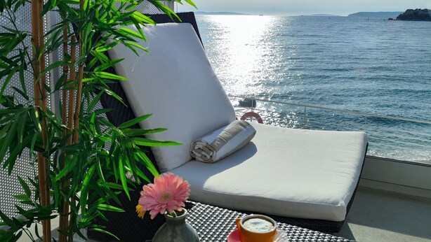 A sunlit balcony with a white cushioned lounge chair, a potted plant, a small table with a drink, and a view of the sea. - Hotel Split (Podstrana)