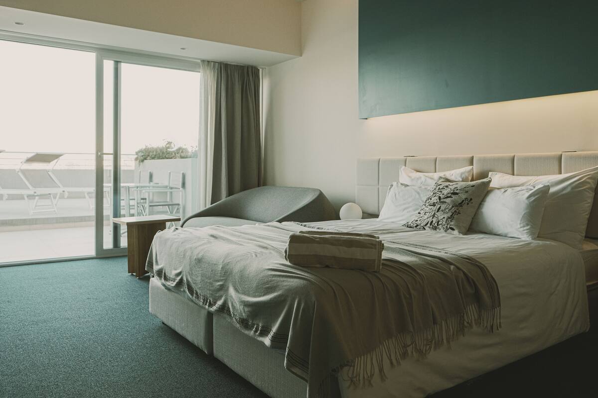 A modern hotel room with a large bed, a bedside table, a lamp, and a view of the outdoors.