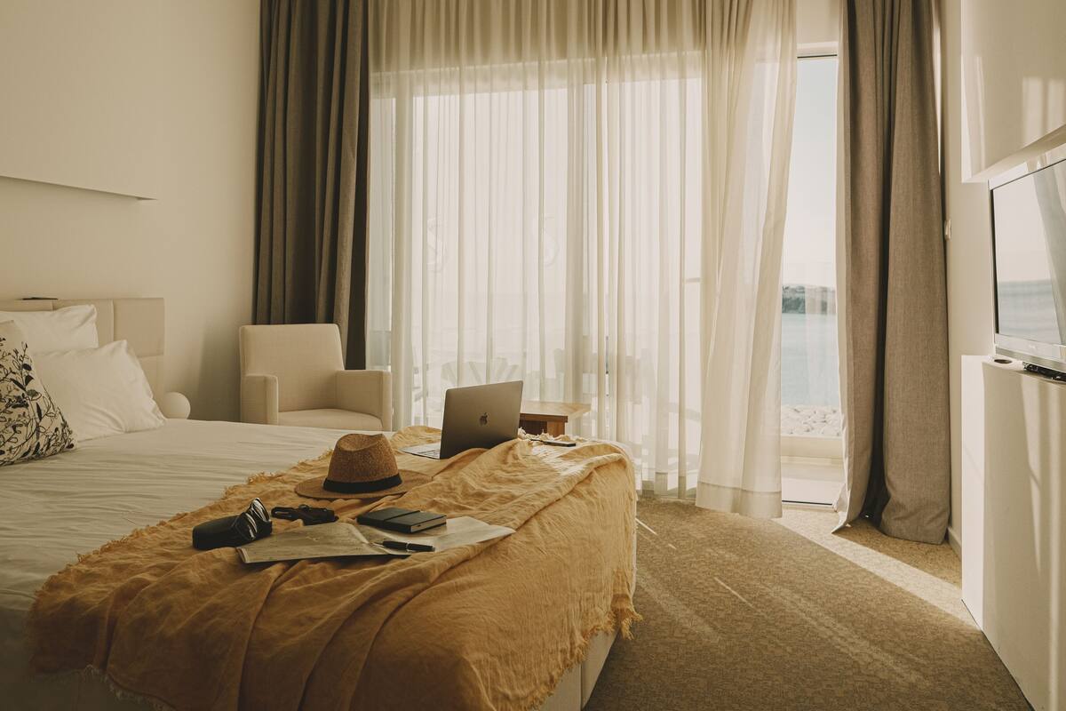 A hotel room with a bed, a laptop on a small table, a hat, and a blanket.
