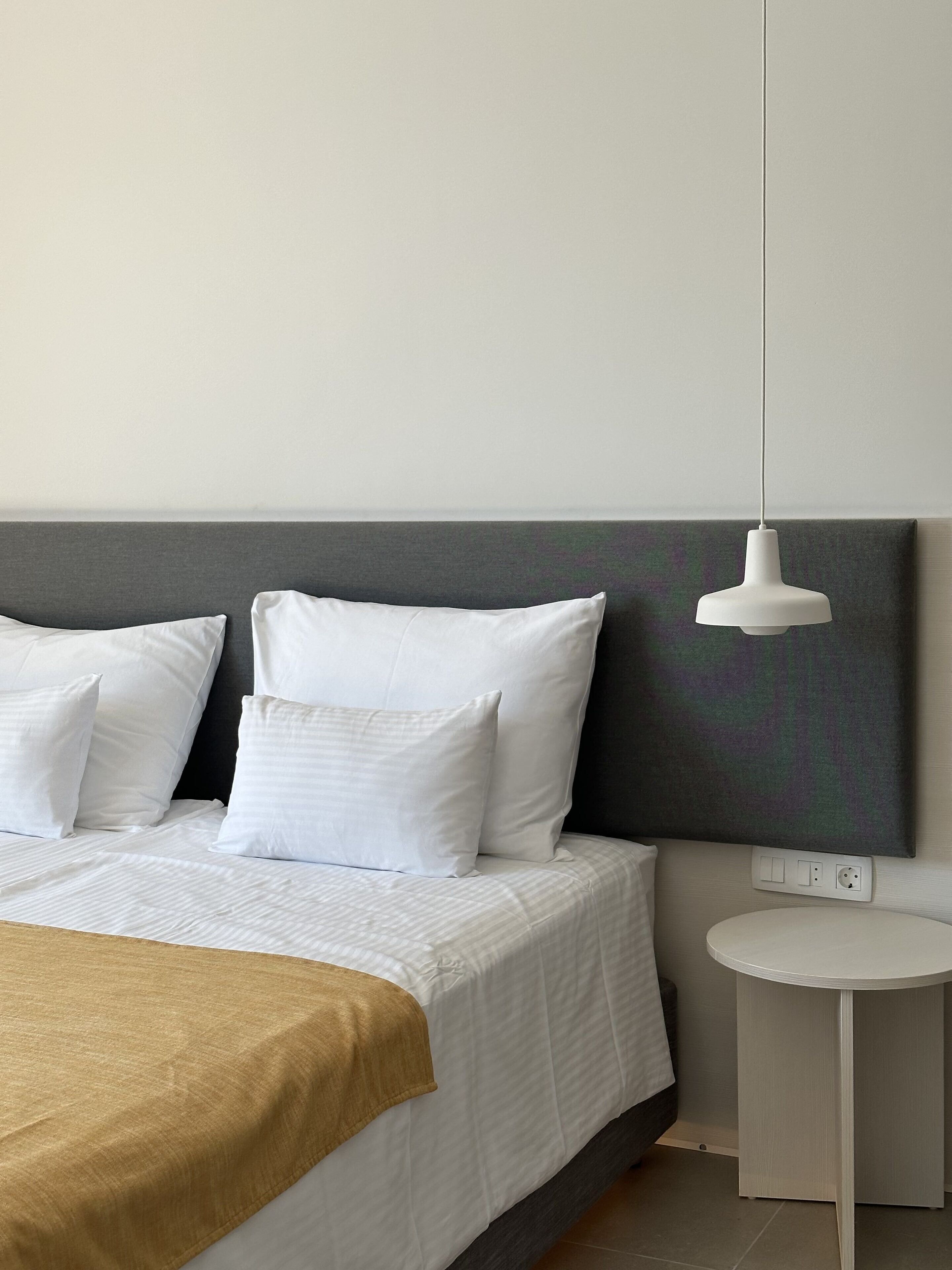 A modern bedroom with a bed, white pillows, a grey headboard, a hanging pendant light, and a small bedside table.