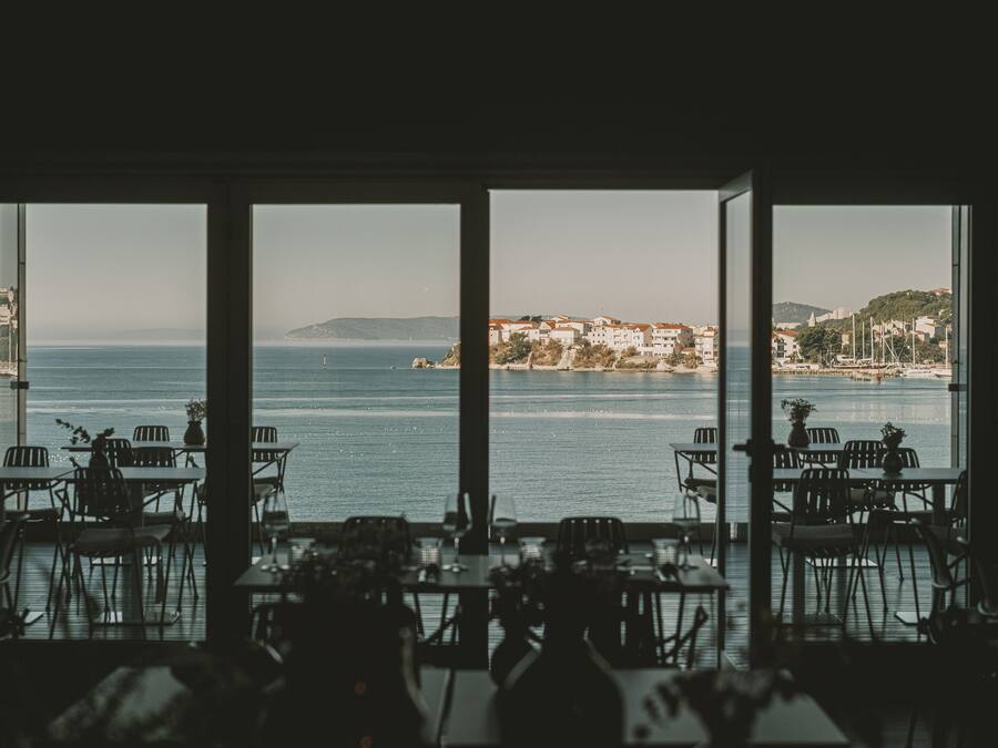 A restaurant with large windows overlooking a coastal view, featuring tables and chairs arranged for dining.