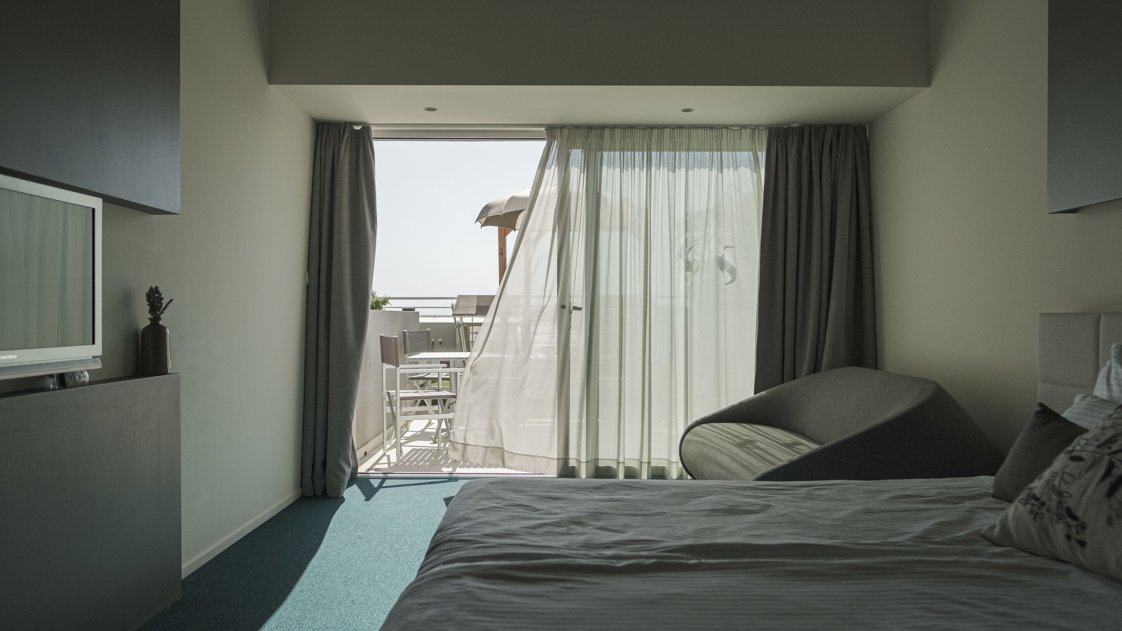 Habitación de hotel con cama, una silla, televisor y balcón con cortinas.