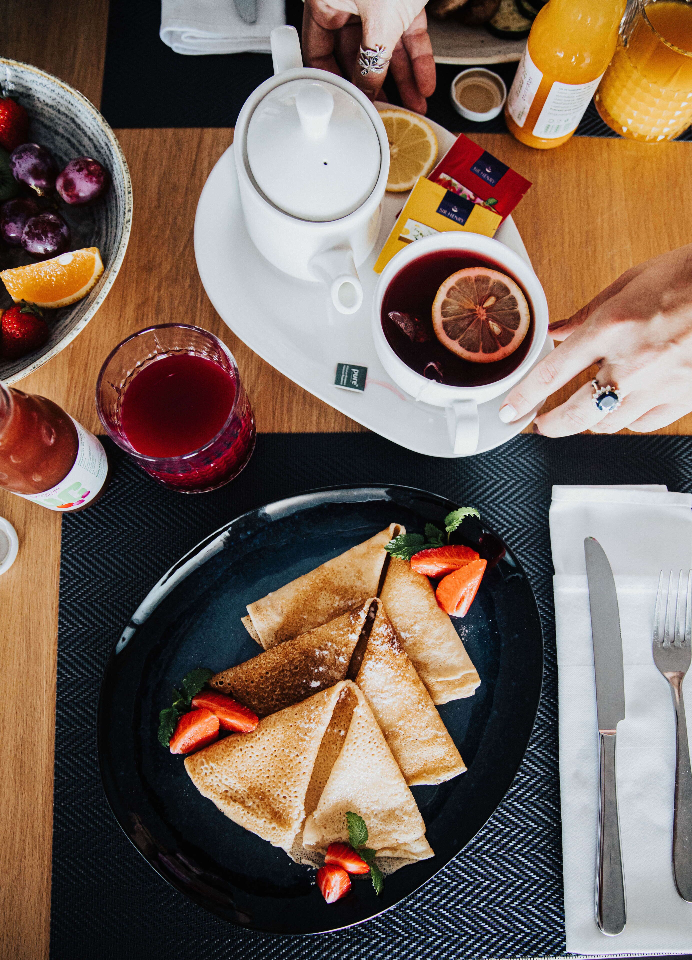 Una mesa con crepas, una taza de té y un vaso de jugo de frutas rojas.
