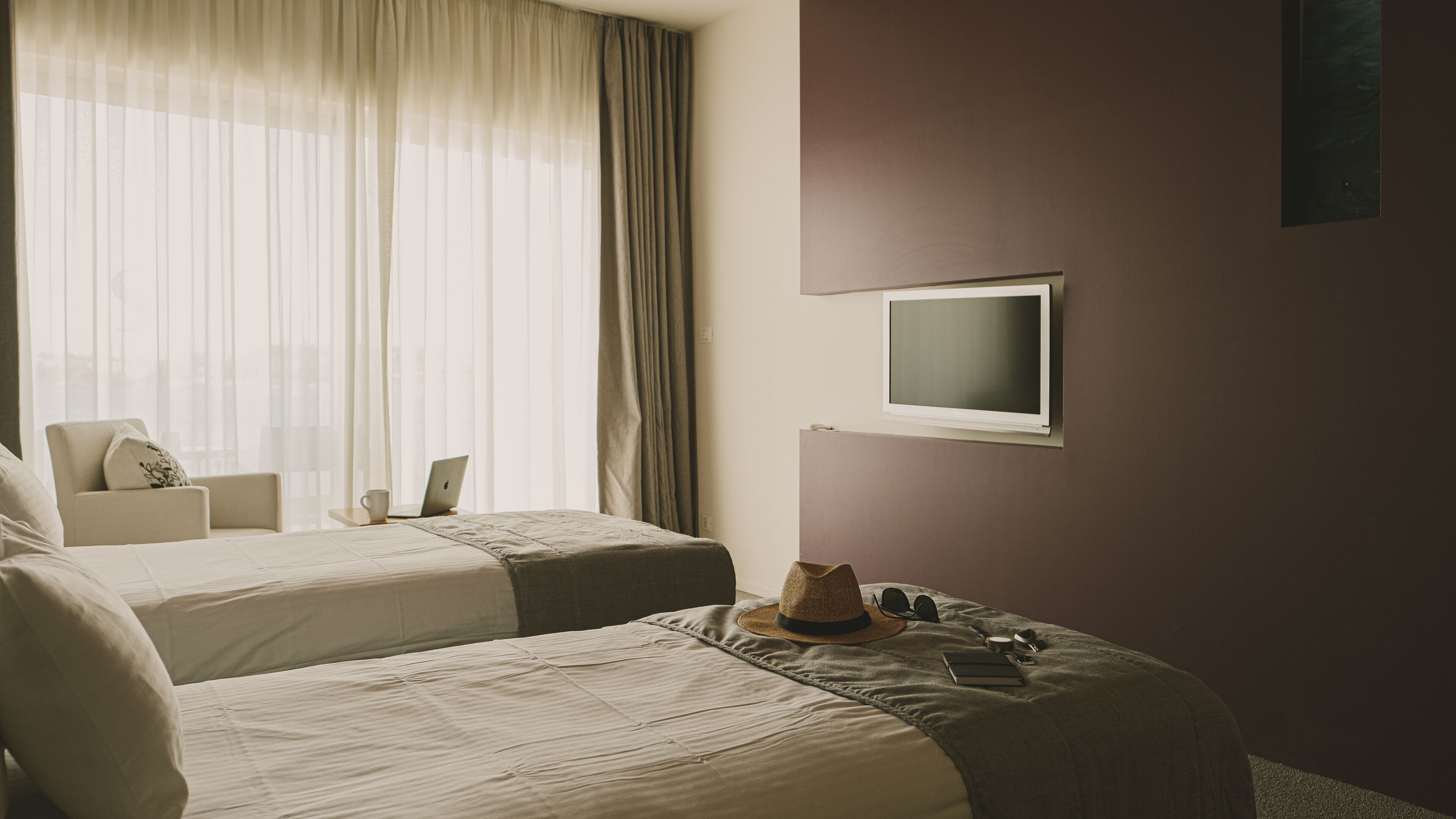 A hotel room with two beds, a desk with a laptop, a TV mounted on the wall, and a large window with sheer curtains.