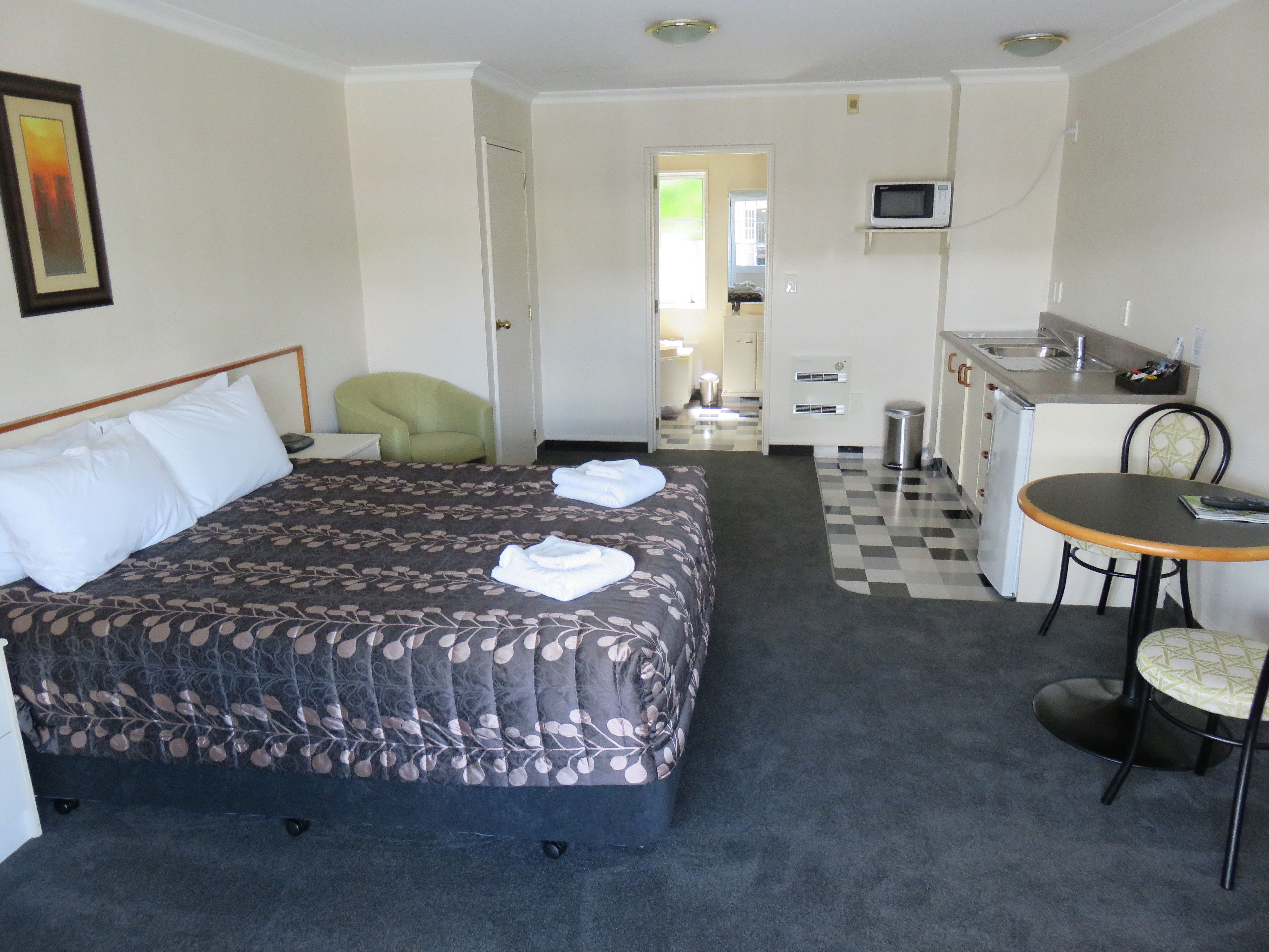 A hotel room with a bed, a green armchair, a small kitchenette, and a dining area.