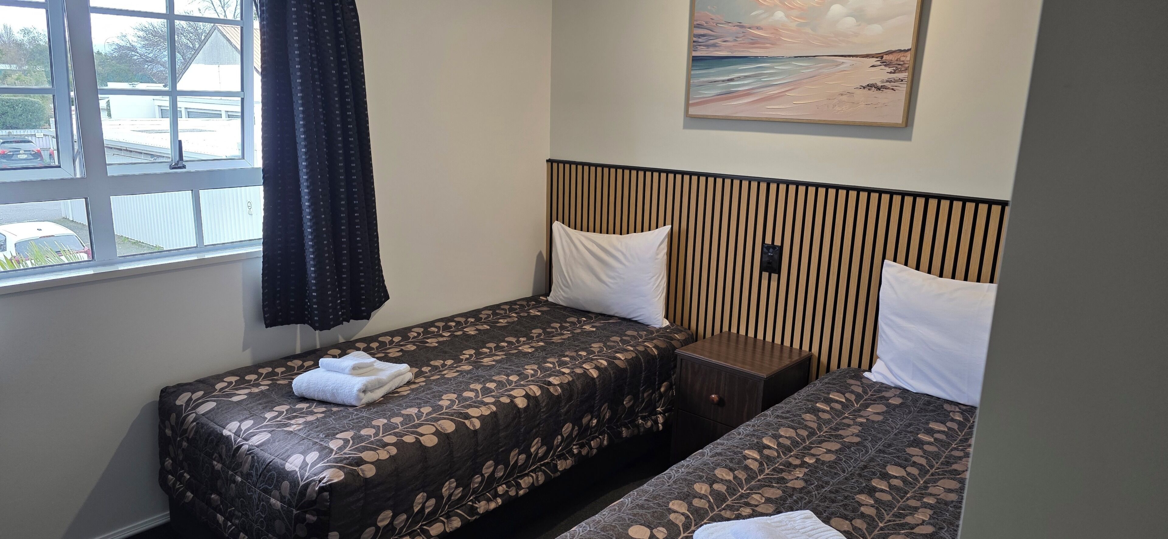 A hotel room with two beds, a window with curtains, a painting on the wall, and a small bedside table.