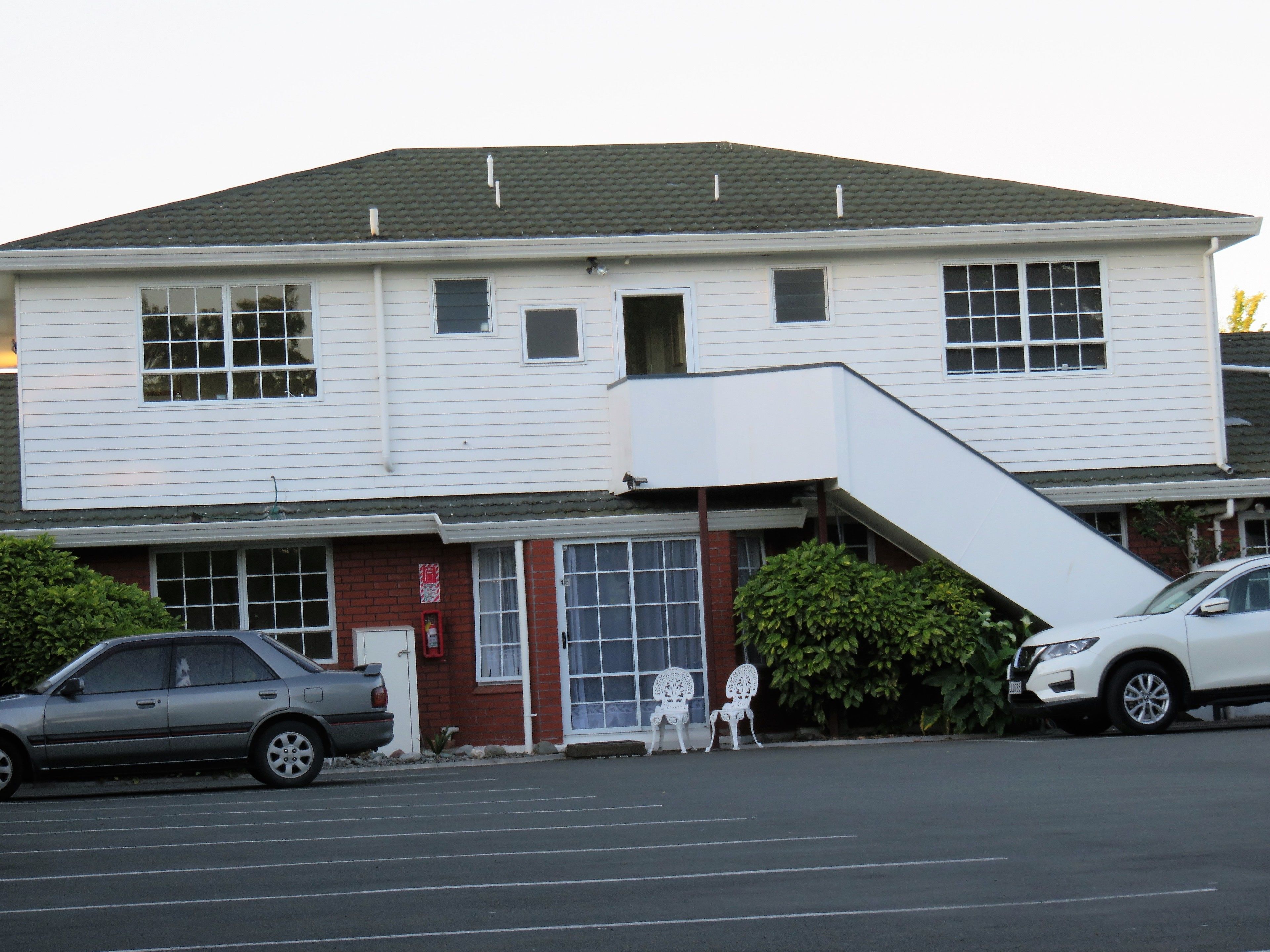 A two-story building with a flat roof and a front entrance with a small porch. There are two cars parked in front, and a white chair is placed outside the entrance.