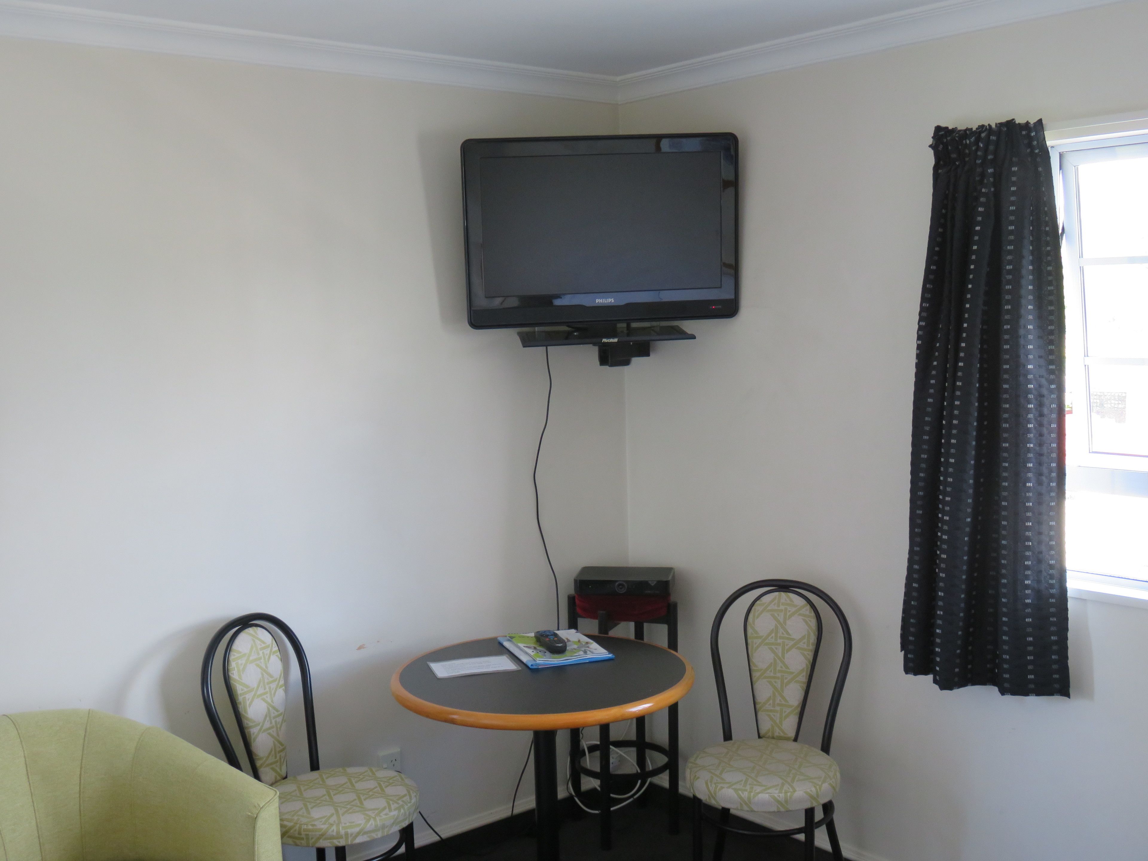 Ein kleiner Raum mit einem an der Wand befestigten Fernseher, einem runden Tisch mit zwei Stühlen und einem Fenster mit Vorhängen.