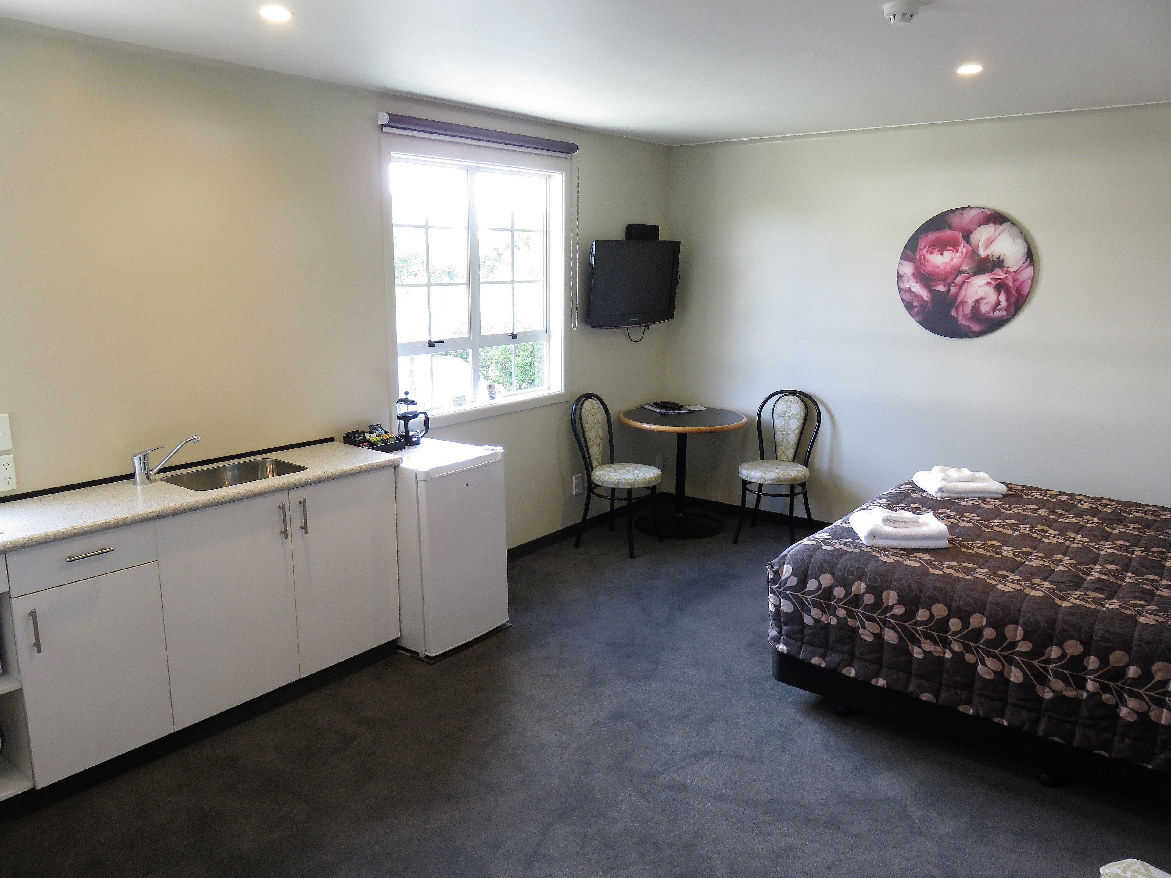 Quarto de hotel compacto com cozinha compacta, uma cama, uma mesinha com duas cadeiras, televisão e um ornamento floral na parede.